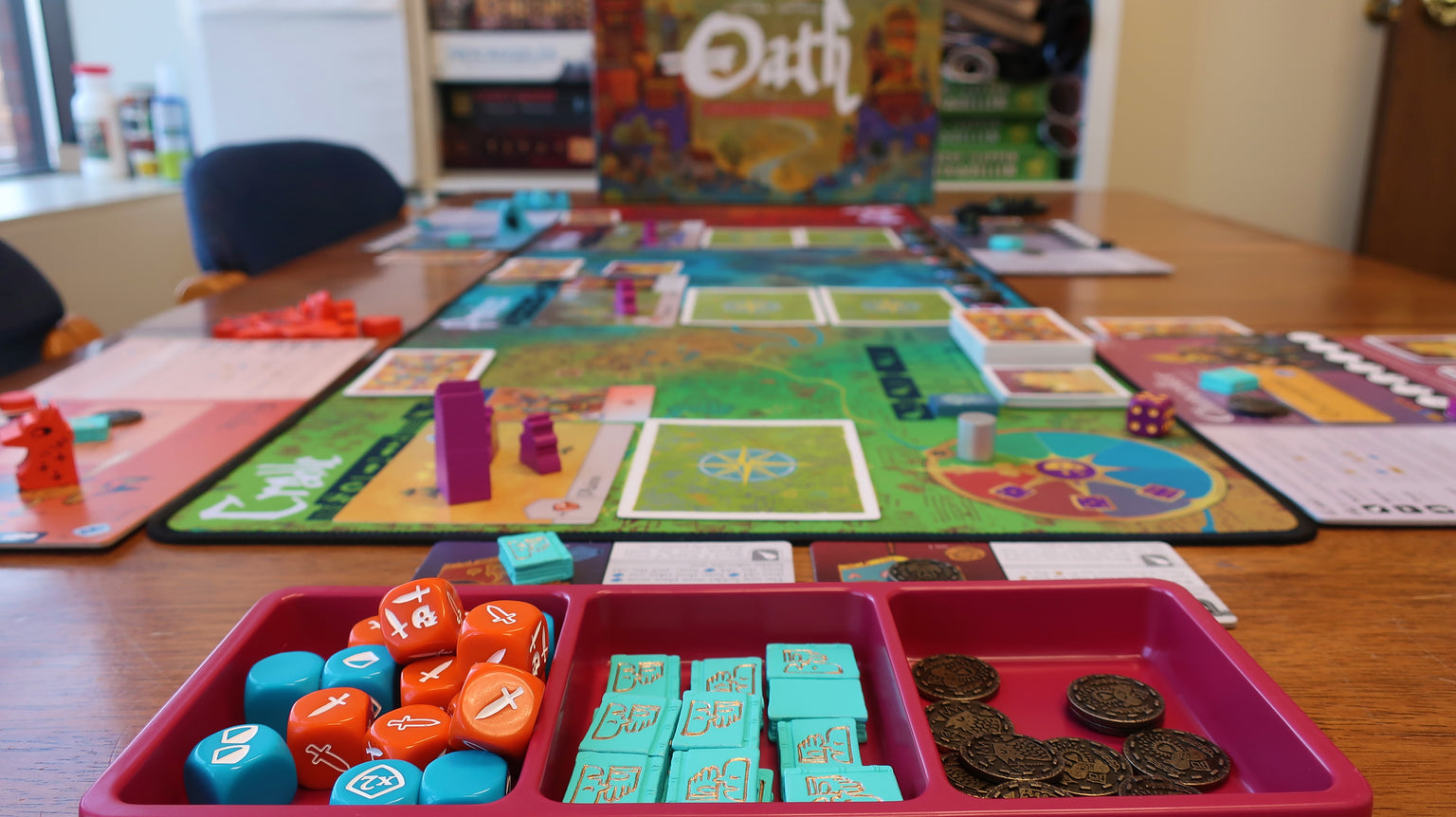 Oath the Board Game set up on a wooden table. Your view is from the far end over a tray with dice and tokens, with the set up board between you and the box cover at the other end.