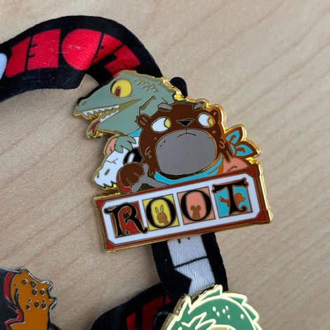 An enamel pin featuring the Root logo as well as character art for the otters and lizards. The pin is on a lanyard.